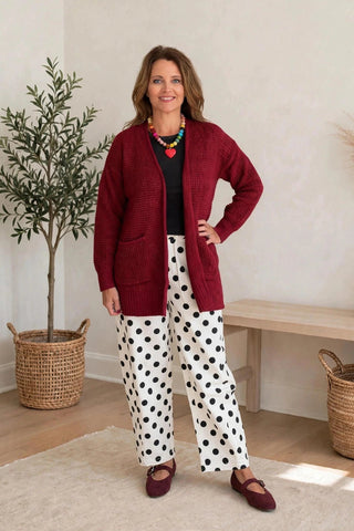 A woman modeling a burgundy cardigan over a black top and white pants with black polka dots, standing in a brightly lit room with a potted plant and a bench in the background.