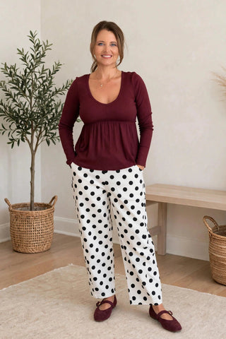 A woman modeling a two-piece loungewear set consisting of a burgundy long-sleeve peplum top and white pants with black polka dots, standing in a brightly lit room with a potted plant and wooden bench in the background.