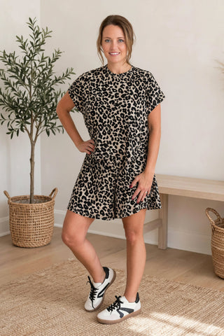 A woman modeling a short-sleeve black and beige leopard print romper dress, standing in a bright room with a potted plant, wooden bench, and woven baskets.