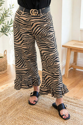 A person wearing wide-leg cropped pants with a zebra print pattern in beige and black, styled with a black belt featuring a gold double-G buckle and black slide sandals, standing on a woven rug in a bright room.