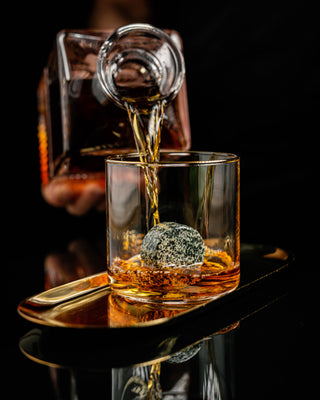Whiskey being poured from a decanter into a glass with a decorative rock on a dark background