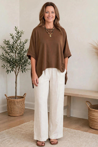 A woman modeling a brown short-sleeve top and white wide-leg pants in a bright indoor setting with a potted plant, bench, and woven baskets.